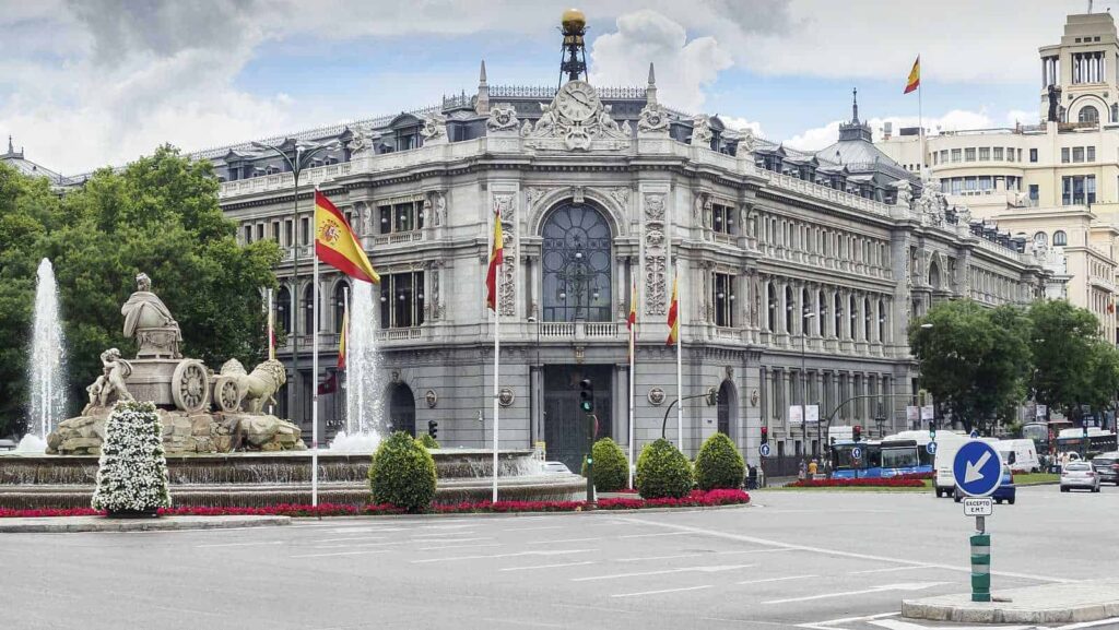 Vista exterior del Banco de España, un edificio monumental en la Plaza de Cibeles, con banderas españolas ondeando y fuentes en el primer plano.