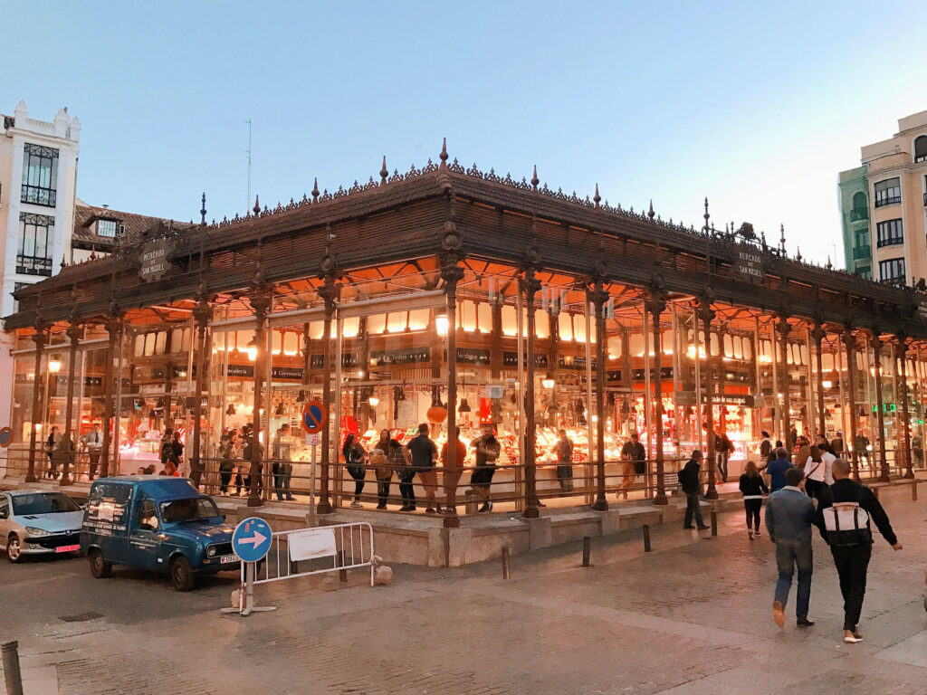 Vista exterior del Mercado de San Miguel en Madrid, con su distintiva arquitectura de hierro y gente caminando frente a la entrada iluminada.
