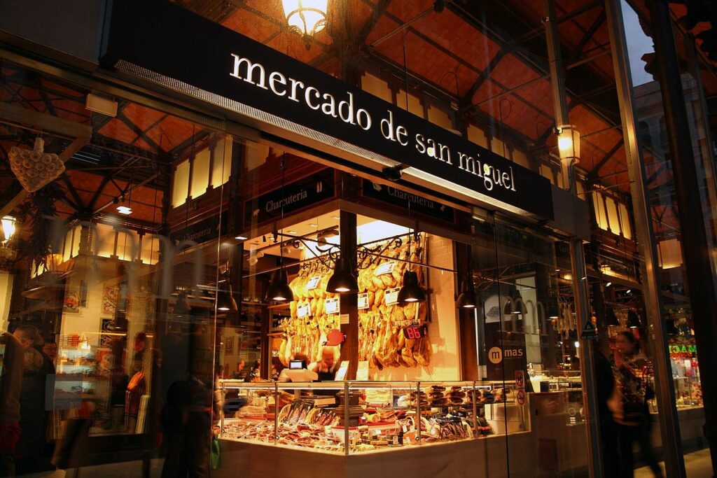 Vista del puesto de charcutería en el Mercado de San Miguel, con productos expuestos y el letrero del mercado iluminado.