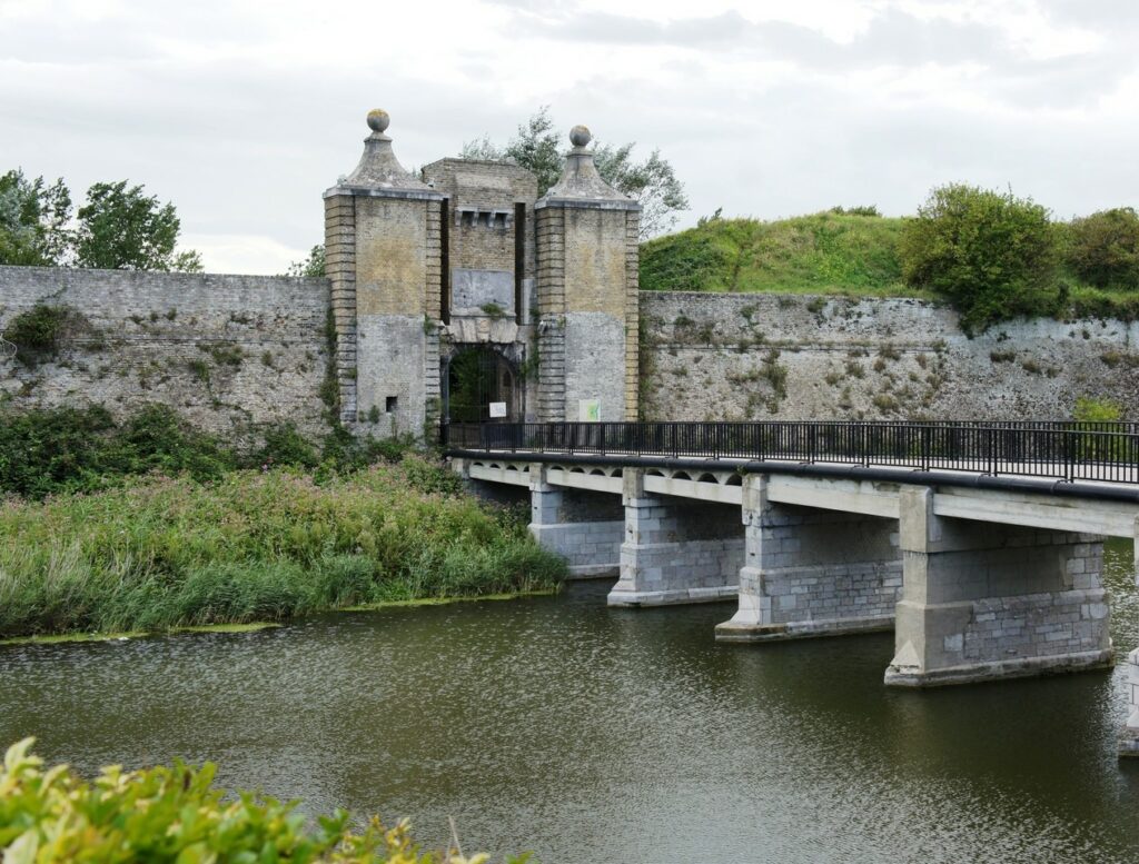 Imagen de un antiguo puente de piedra que conecta dos lados de un río, con una estructura histórica y un muro de fortificación en el fondo, rodeada de vegetación.