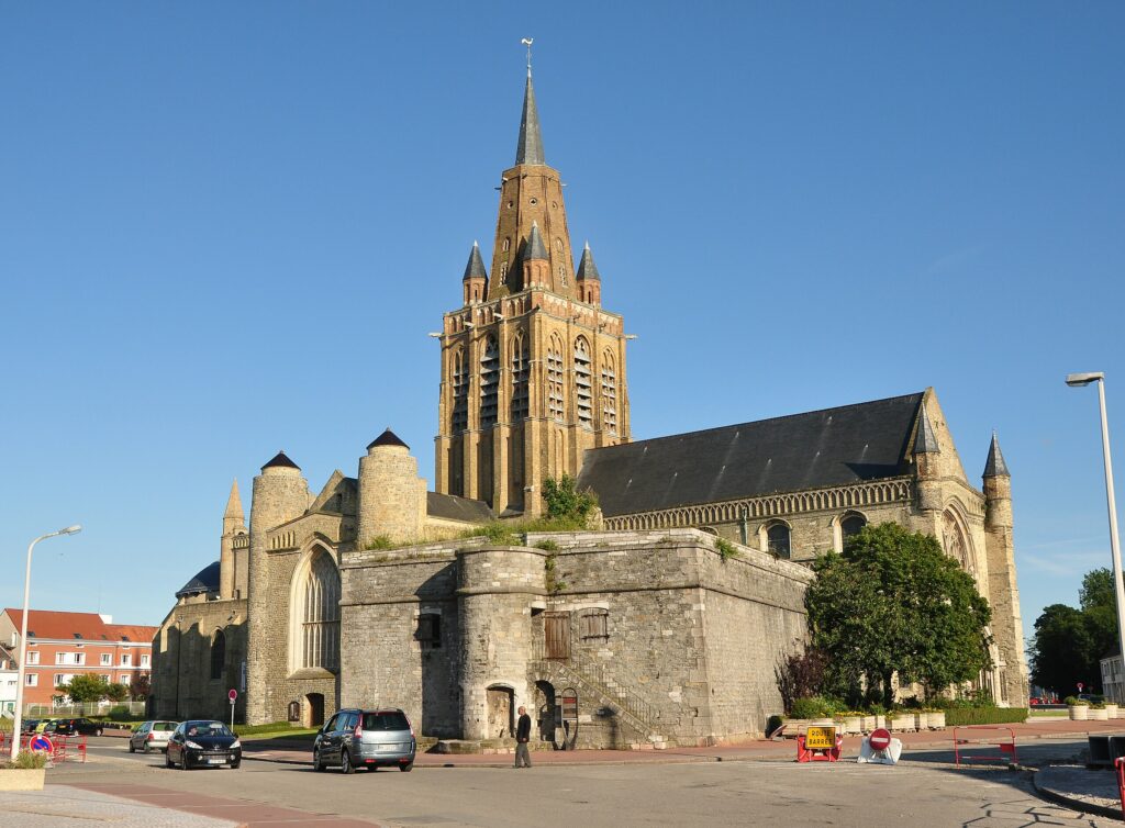 Imagen de la iglesia de Notre-Dame en Calais, Francia, con su distintiva torre y arquitectura histórica, rodeada de vehículos y vegetación.