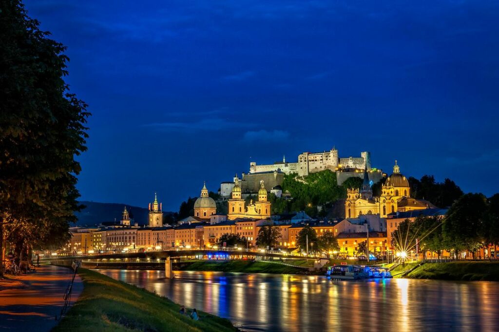 Vista nocturna de Salzburgo, Austria, mostrando la fortaleza Hohensalzburg sobre la ciudad iluminada y el río Salzach reflejando las luces.