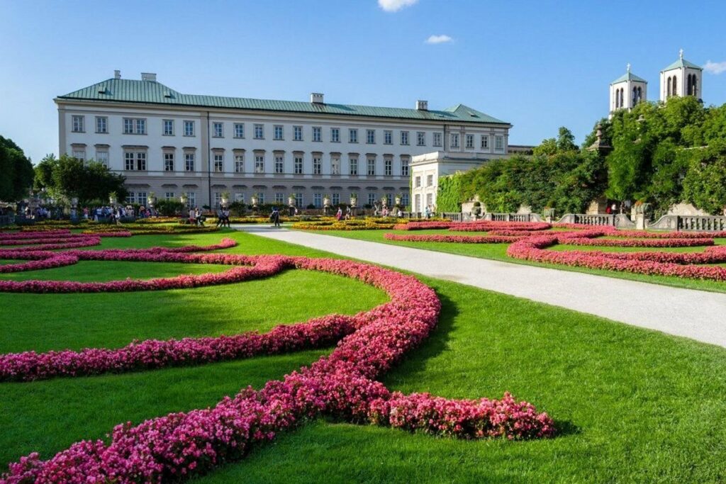 Jardines del Palacio de Mirabell en Salzburgo, Austria, con diseño florido y césped verde, rodeados de personas disfrutando del entorno.