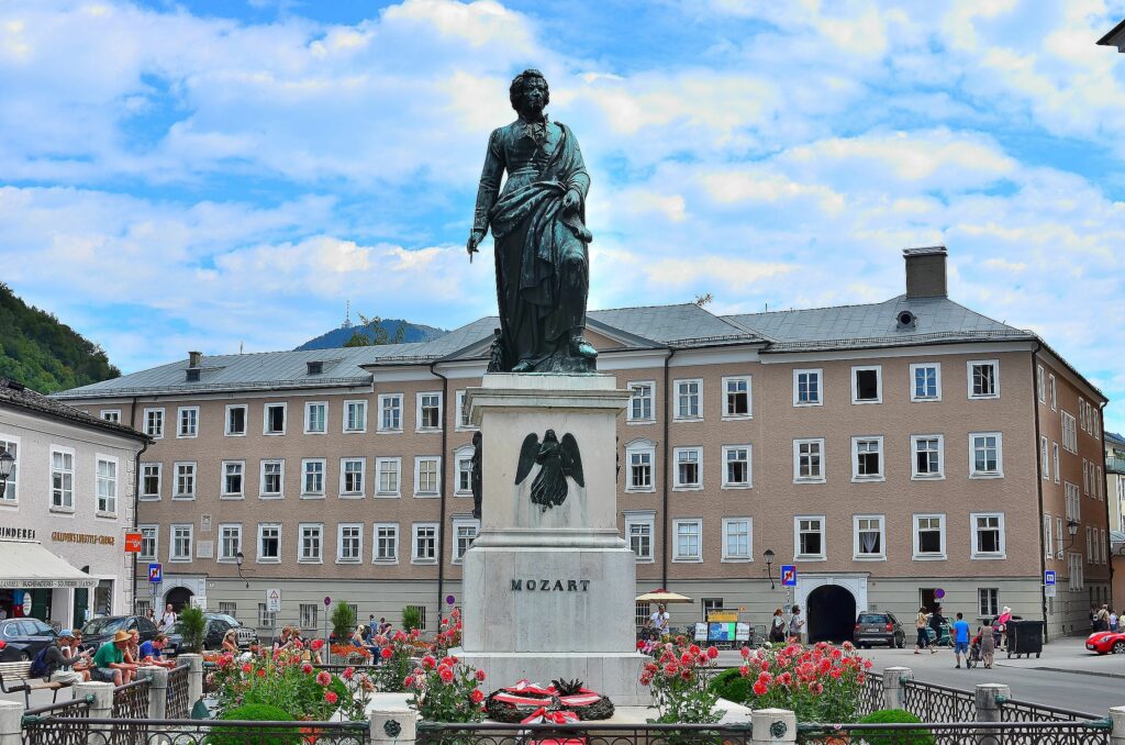 Estatua de Mozart en una plaza de Salzburgo, rodeada de flores y edificios históricos bajo un cielo azul.